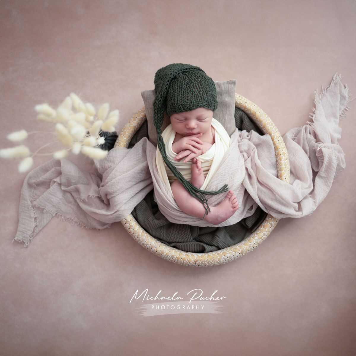 Schlafendes Neugeborenes sitzt eingewickelt in helle Tücher in einem runden Korb; es trägt eine dunkelgrüne Strickhaube und ist umgeben von weichen Stoffen und zarten Naturakzenten.